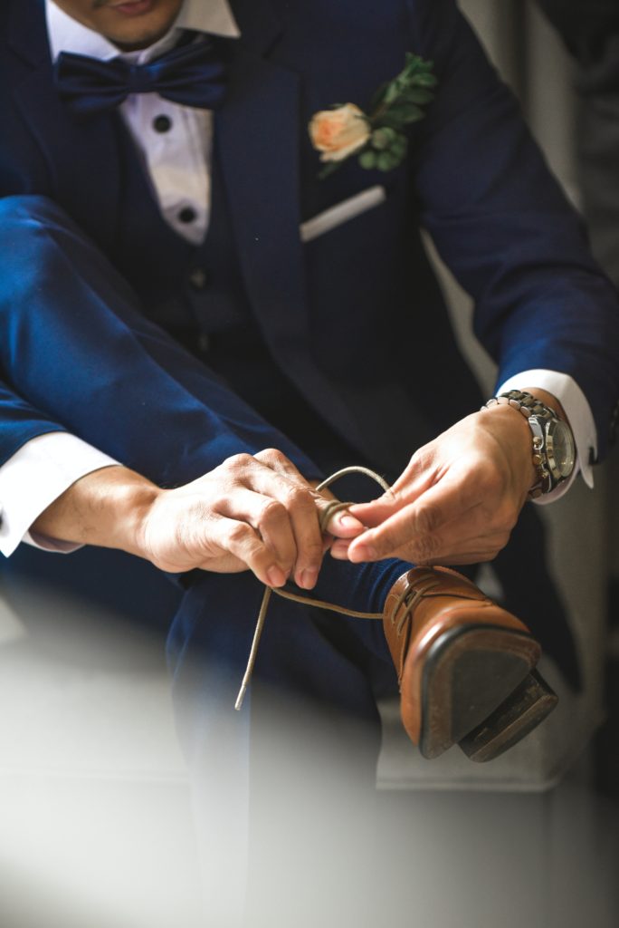 Groom ties his shoes on wedding day wearing a navy suit and brown shoes